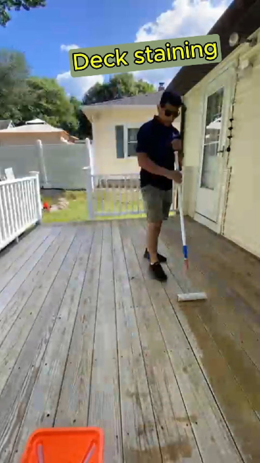 Person staining a weathered wooden deck beside a house, with a roller and paint tray.