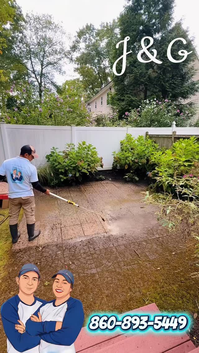 Man spraying a patio with a pressure washer, clearing mud and debris near shrubs and a fence.