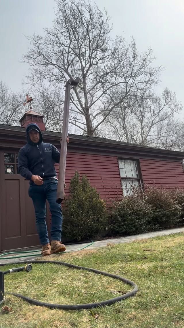 Person in hoodie standing on lawn with a hose in front of a red house and leafless tree