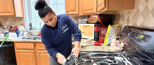 Person cleaning a kitchen stove beside wooden cabinets and a cluttered countertop