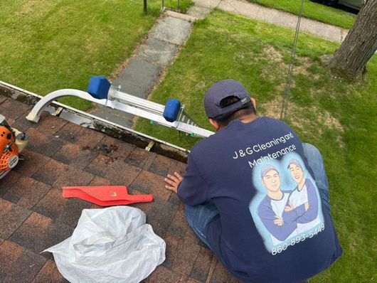 Person on a roof wearing a navy shirt, near a ladder, tools, and a white bag by a yard walkway.