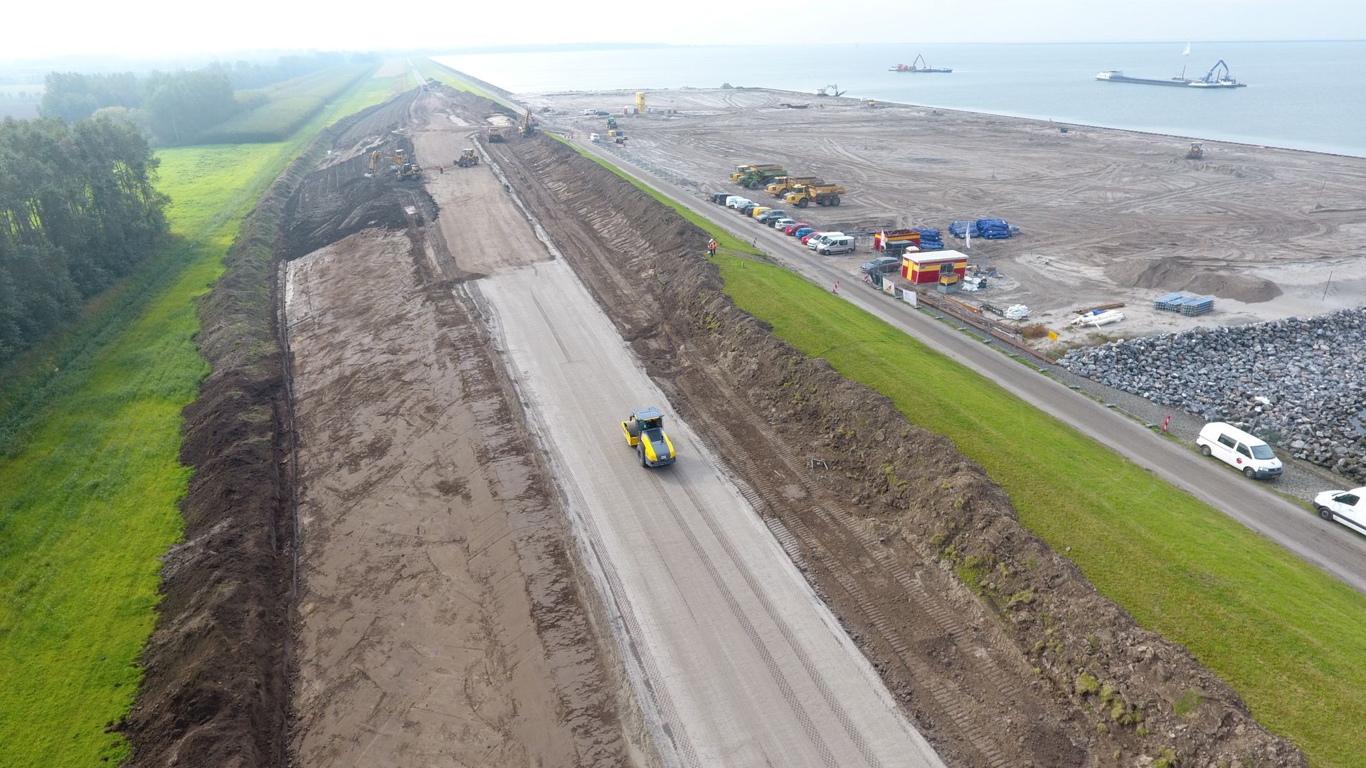 Luchtfoto van bouwwerkzaamheden aan een dijk langs een waterlichaam, met zwaar materieel en een gele wals op de weg.