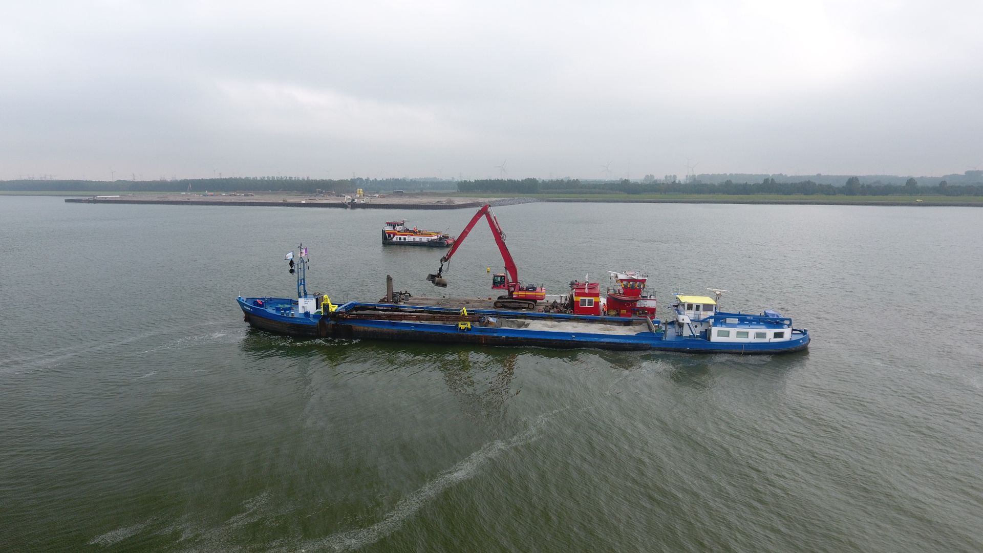 Een blauw werkschip met een rode kraan ligt op kalm water, met in de verte, onder een bewolkte hemel, nog een boot zichtbaar.