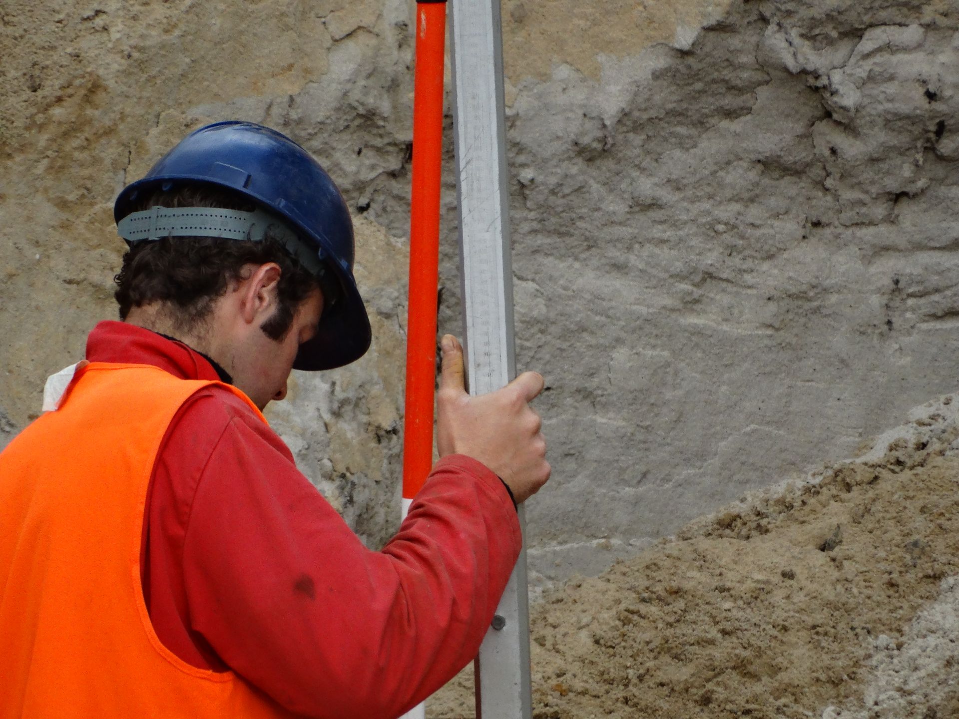Een arbeider in een rood shirt en oranje vest houdt een verticale nivelleerstok vast op een bouwterrein met aarden muren.