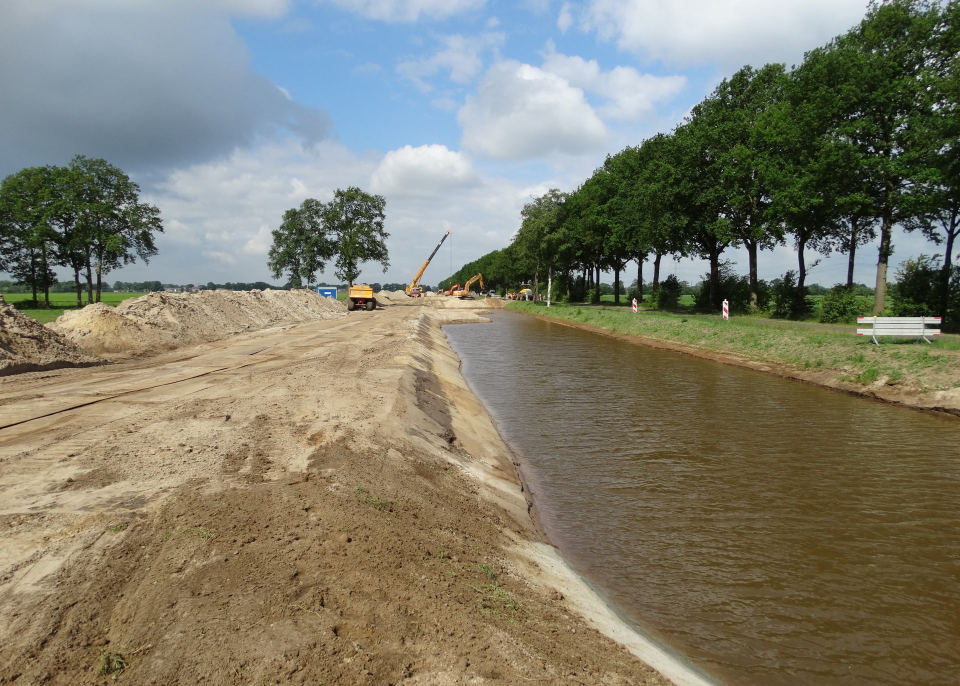 Een bouwterrein langs een waterweg, met hopen grond, zwaar materieel in de verte en een rij bomen.