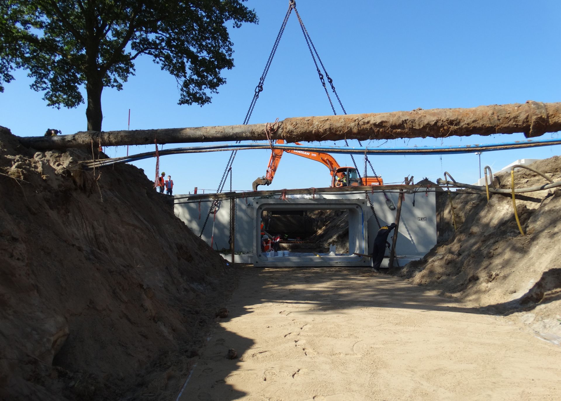 Bouwvakkers plaatsen een grote betonnen kokerduiker in een uitgegraven sleuf onder een hangende nutsleiding.