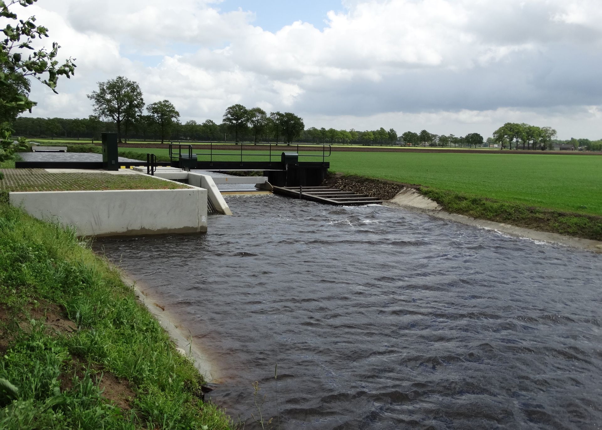 Een kleine betonnen stuw overspant een donker stromend beekje in een grasrijk, open landschap onder een bewolkte hemel.