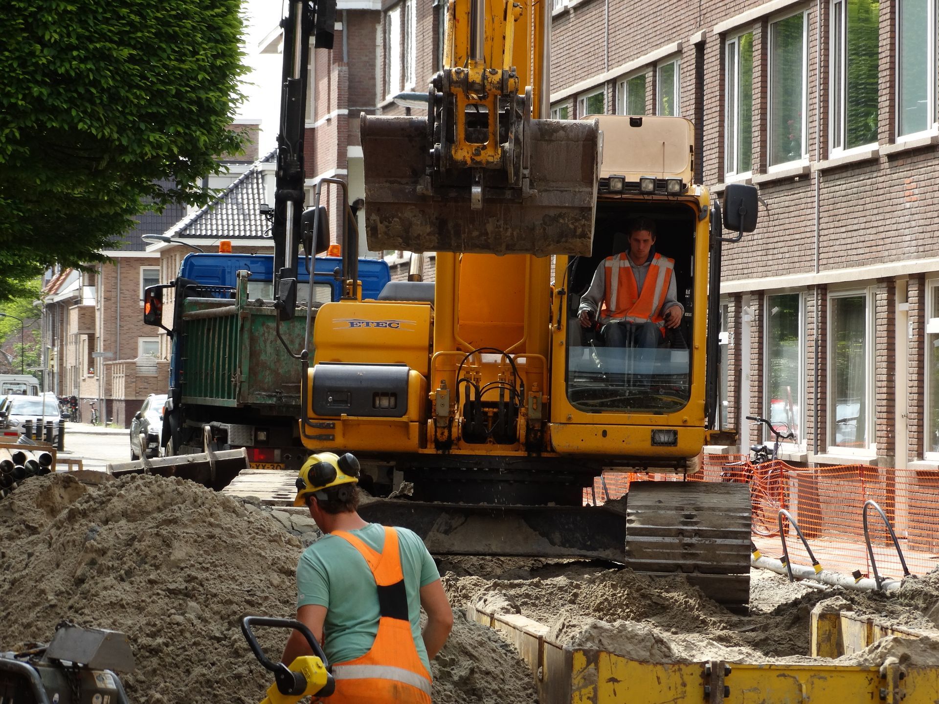 Een bouwvakker staat naast een gele graafmachine die een sleuf graaft in een woonstraat.