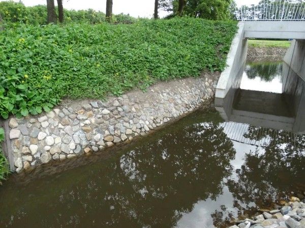 Een betonnen duiker overspant een beek met een stenen keermuur en een met gras begroeide oever.