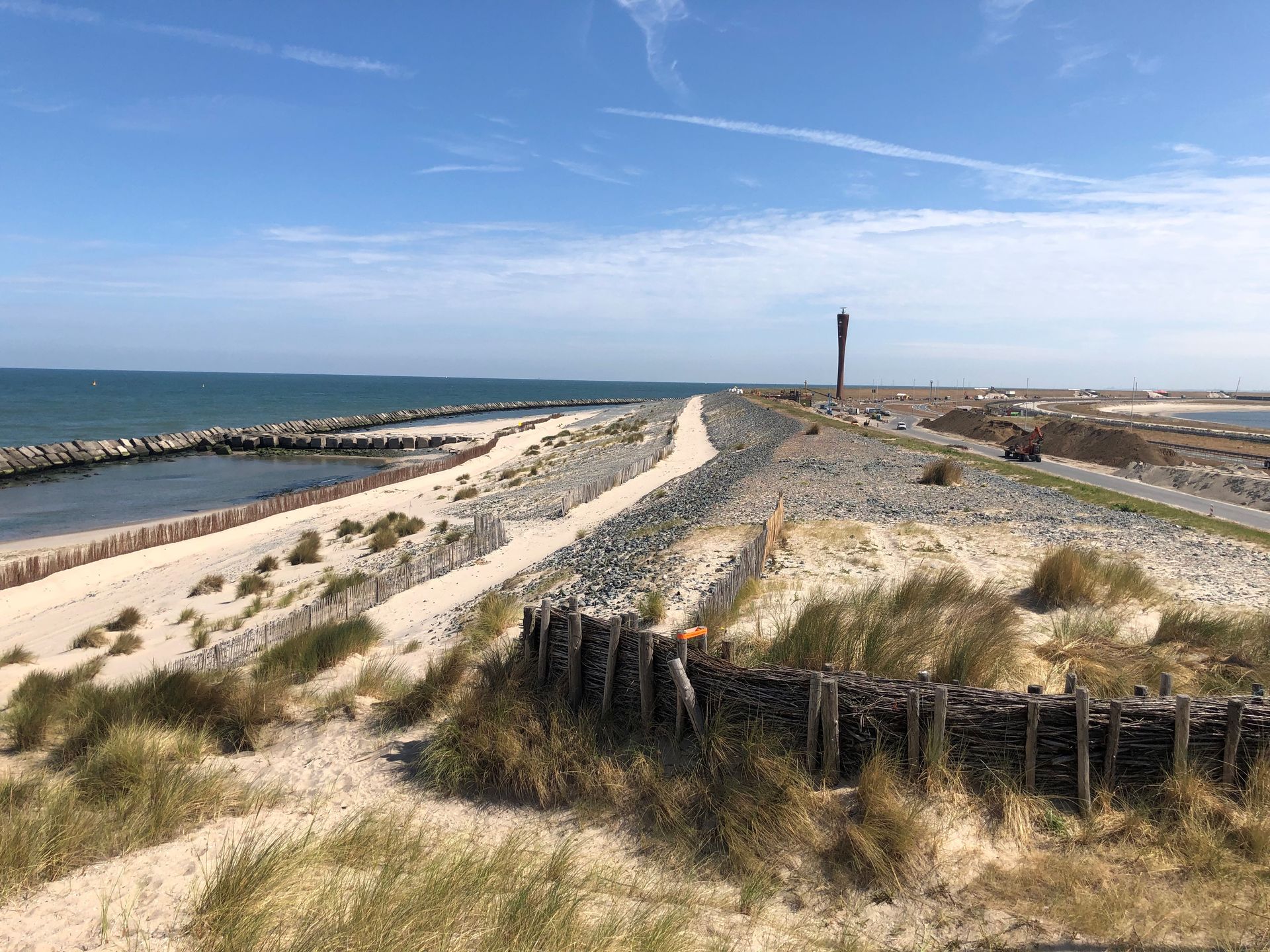 Kustlandschap met een zandpad, duinafscheiding, rotsachtige golfbreker en de oceaan onder een blauwe hemel.