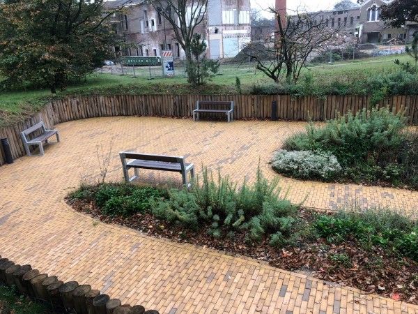 Een betegeld, halvemaanvormig terras met drie banken, omgeven door een houten schutting en aangelegde struiken.