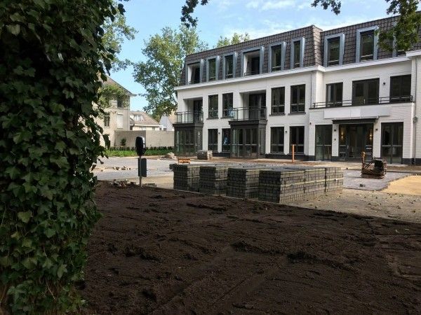 Een nieuw gebouwd wit woonhuis met een donker dak staat achter een pas geëgaliseerd, met aarde bestraat terrein.
