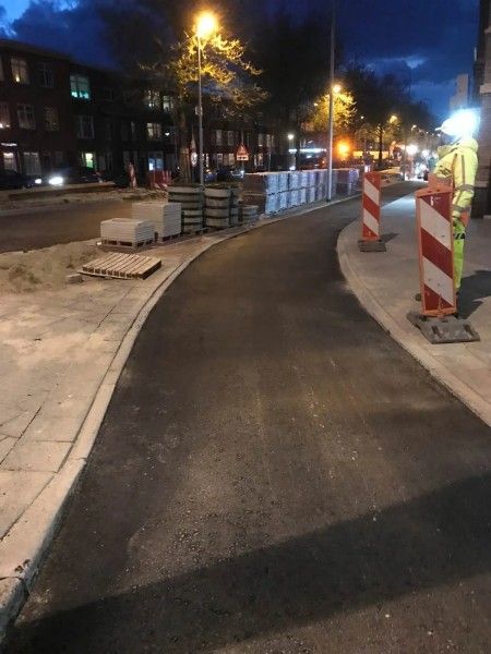 Een pas aangelegd donker asfaltpad 's nachts, begrensd door bouwhekken, straatstenen en straatverlichting.
