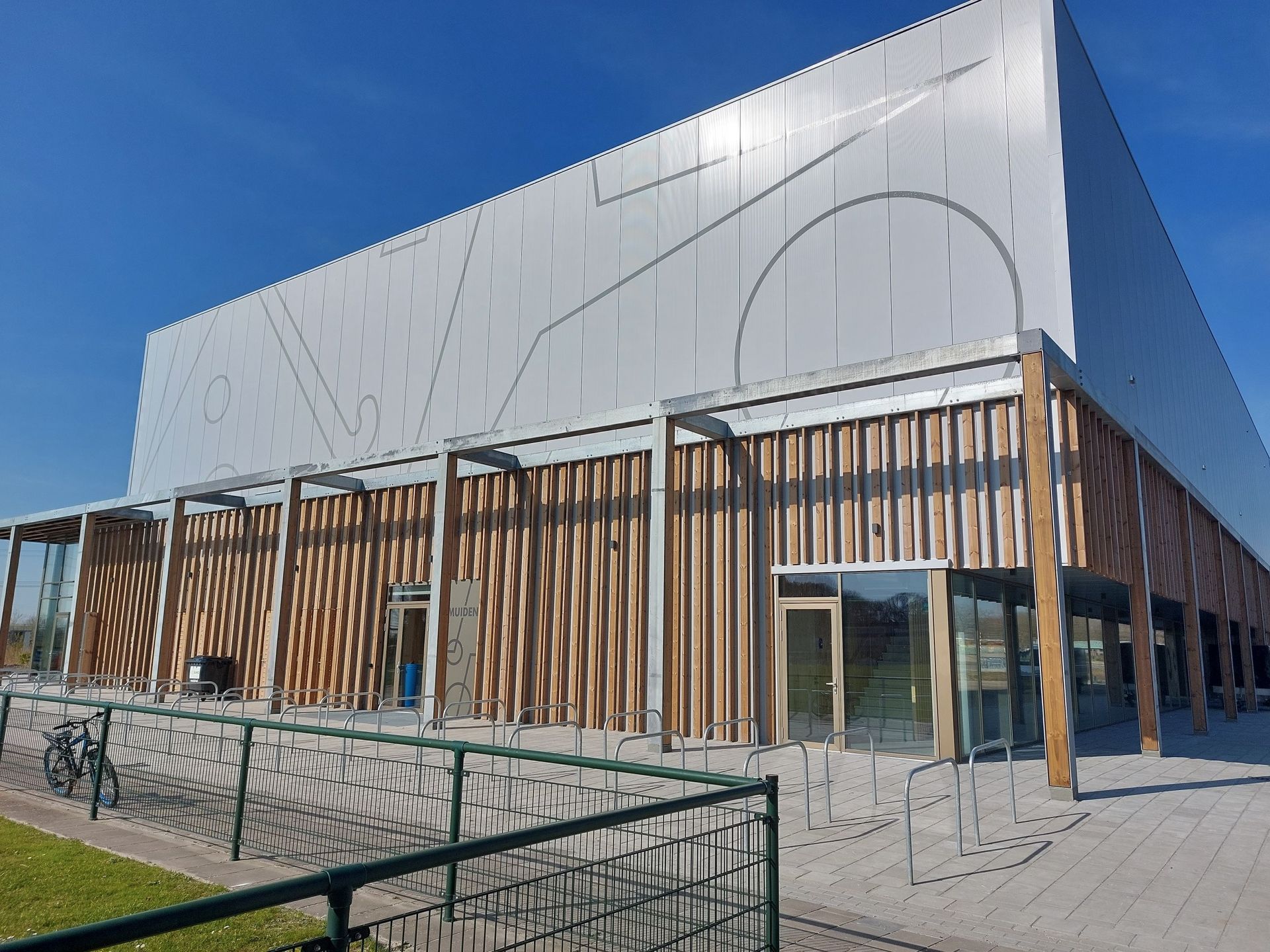 Modern gebouw met een metalen bovengevel en een ondergedeelte met verticale houten latten, vlakbij een fietsenstalling.