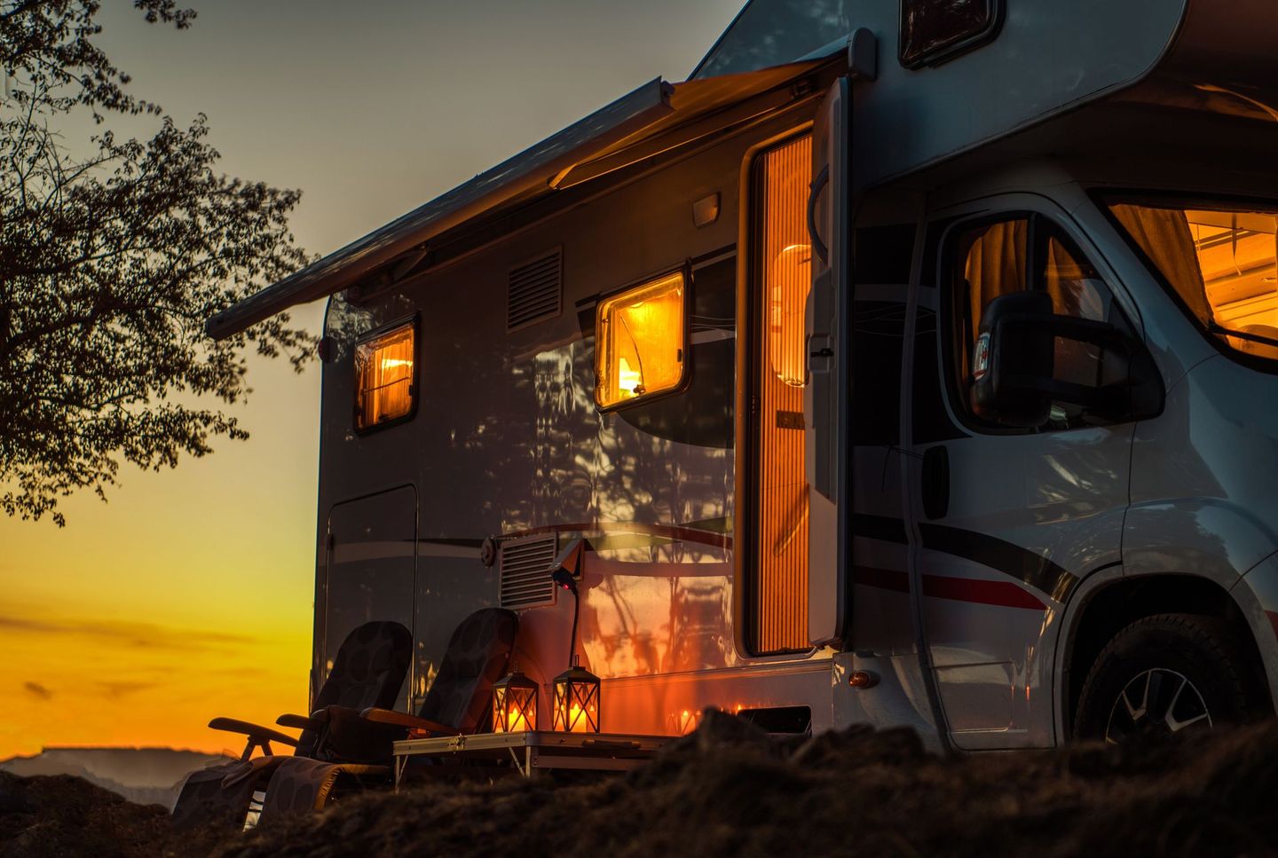 Campervan met gloeiende ramen geparkeerd bij zonsondergang naast bomen en droog gras.