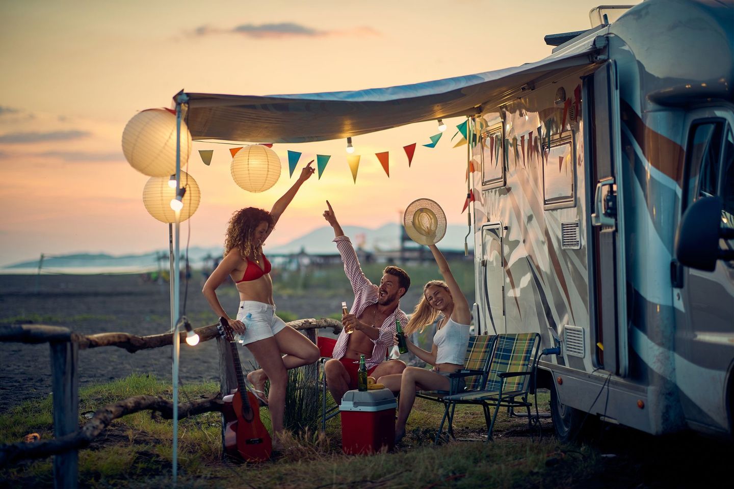 Vrienden picknicken bij een versierde camper bij zonsondergang, met lantaarns, vlaggetjes en trommels op het strand.