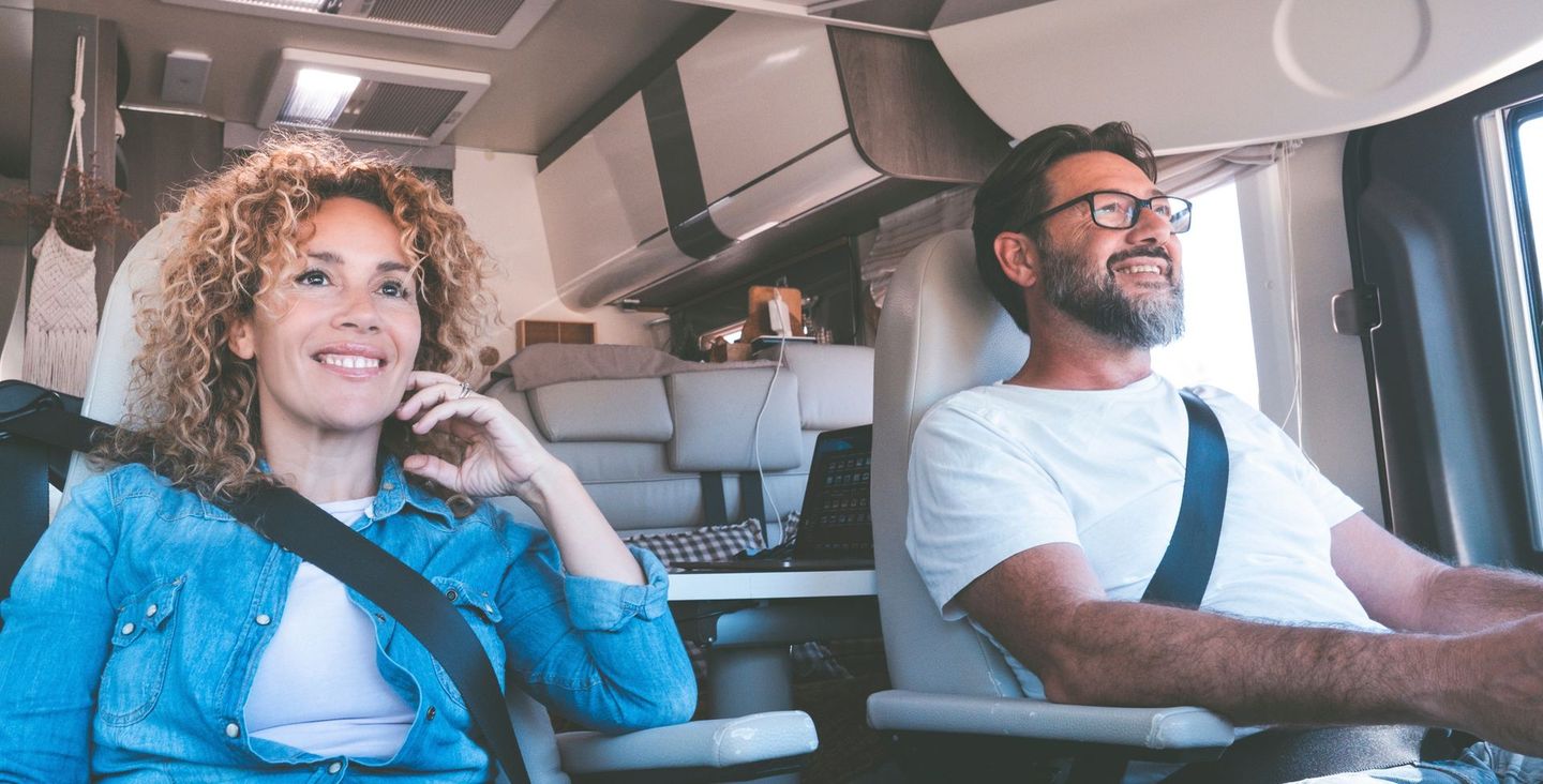 Twee passagiers, glimlachend en vastgesnoerd in een rijdende camper, met de bestuurder en een passagier naast elkaar.