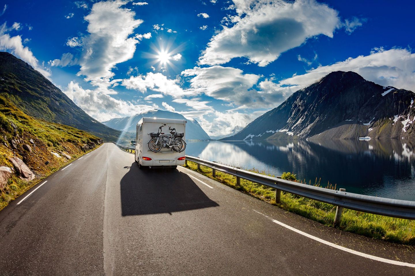 Een witte bestelwagen rijdt over een bergweg langs een rustig meer onder een helderblauwe hemel met wolken.
