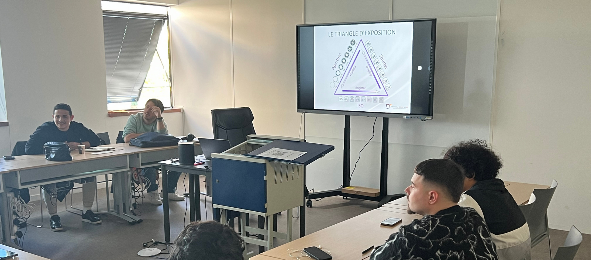 Un groupe de personnes sont assises à des tables dans une salle de classe et regardent une présentation.