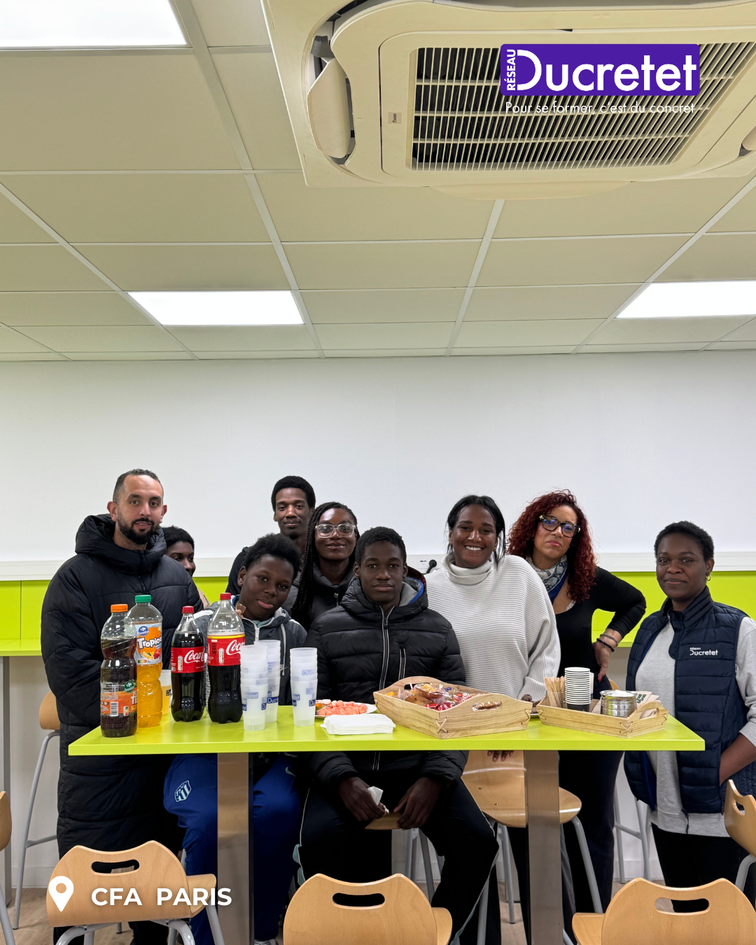Group gathered around a yellow table with drinks and snacks in a bright indoor room, smiling at the camera
