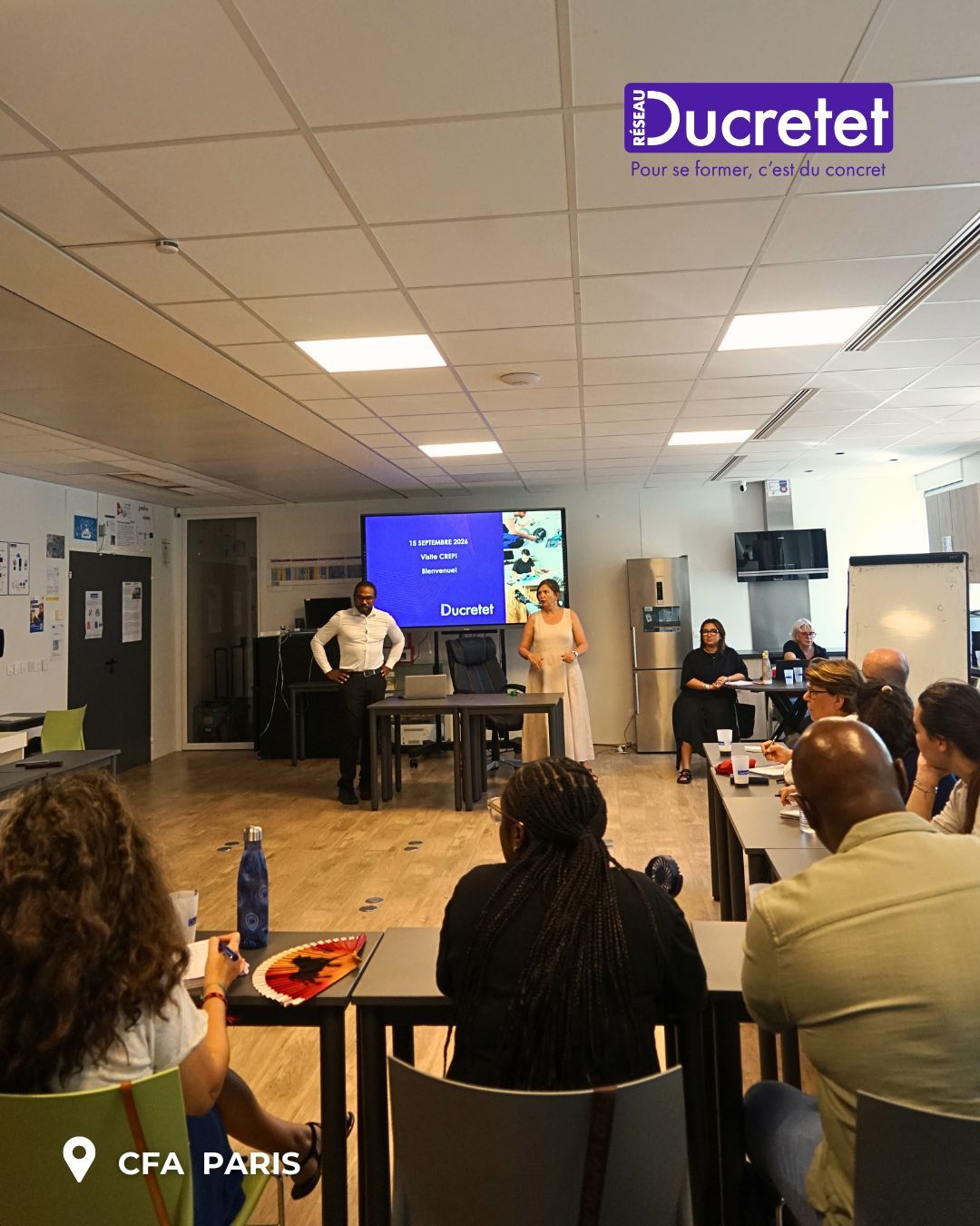 Conference presentation in a classroom, with speaker at the front and audience seated facing a screen.