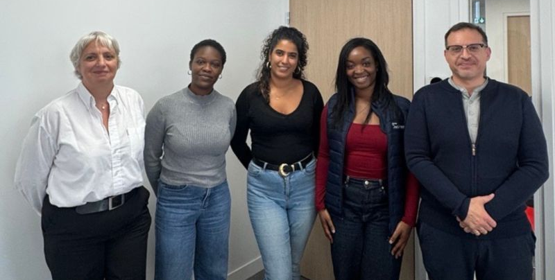Five people stand together indoors, posing in front of a plain wall.