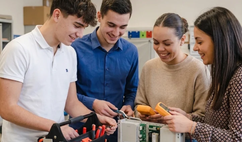 Four students collaborate around electronics components on a lab table, smiling in a classroom.