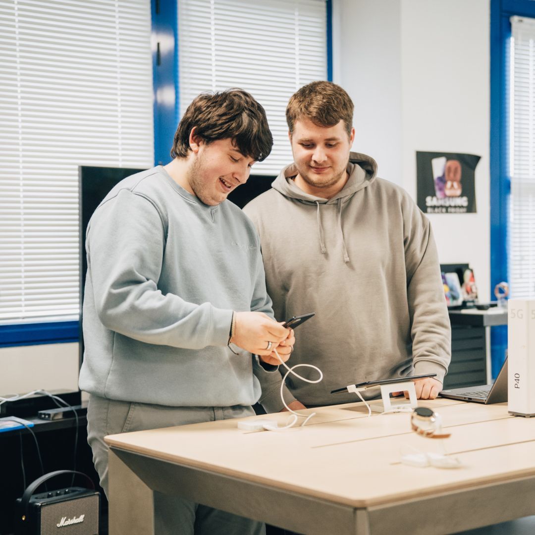 Deux hommes examinent des câbles à une table. L'un sourit, tenant un câble. Pièce claire, fenêtre lumineuse.