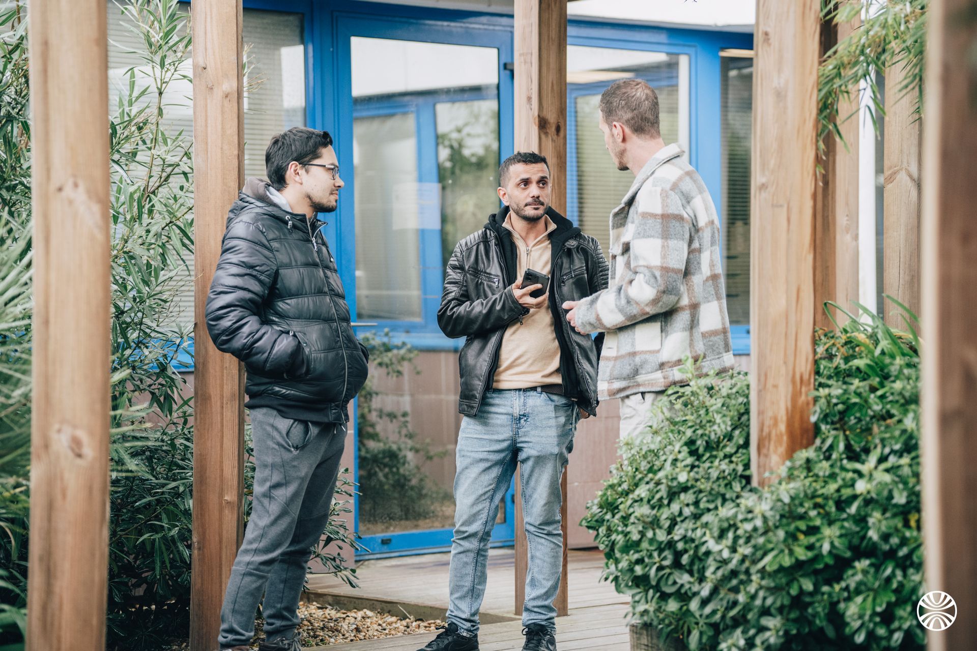 Trois hommes discutent dehors, près de poutres en bois. L'un d'eux tient un téléphone. Ils portent des vestes.