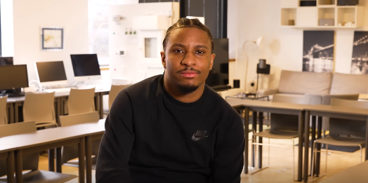 Un homme noir vêtu d'un sweat-shirt noir est assis à l'intérieur, souriant. Une salle de classe se trouve en arrière-plan.