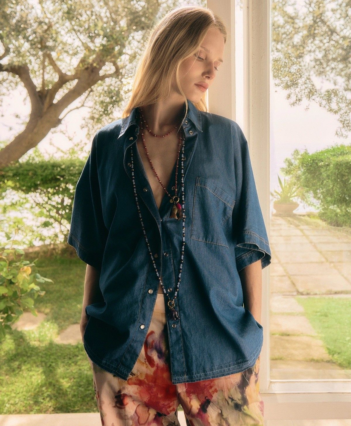 Persona in piedi su una veranda, con indosso una camicia blu larga e pantaloni a fiori, che guarda di lato.