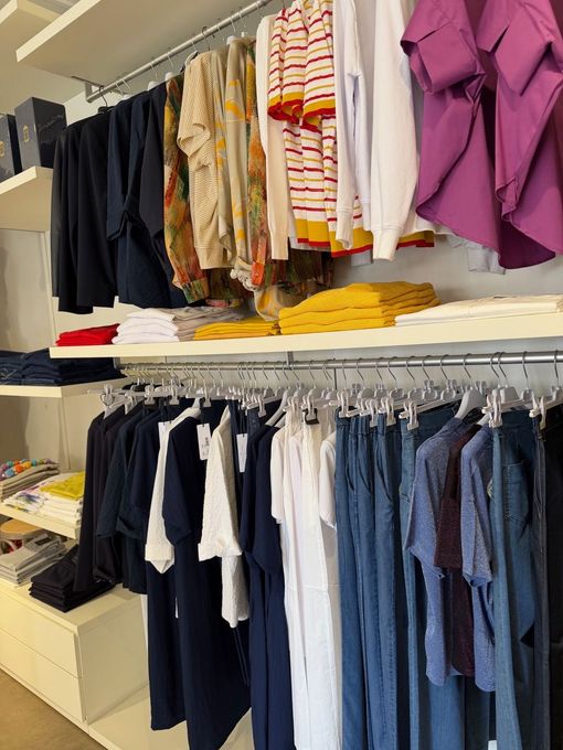 Vetrina di un negozio di abbigliamento con camicie e top appesi nei colori blu navy, bianco, blu, giallo e viola su due appendiabiti.