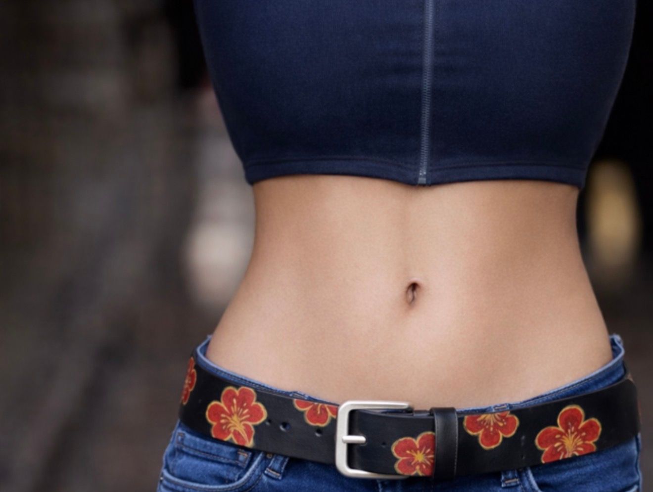 Primo piano di un addome coperto da un top corto scuro e jeans con cintura floreale e fibbia argentata.