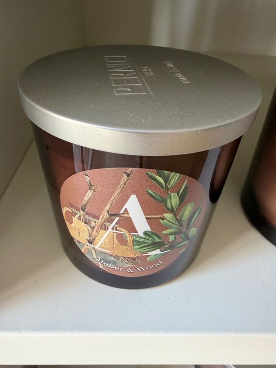 Barattolo portacandele marrone con coperchio argentato su uno scaffale bianco, con etichetta decorata con un motivo di agrumi ed erbe aromatiche.