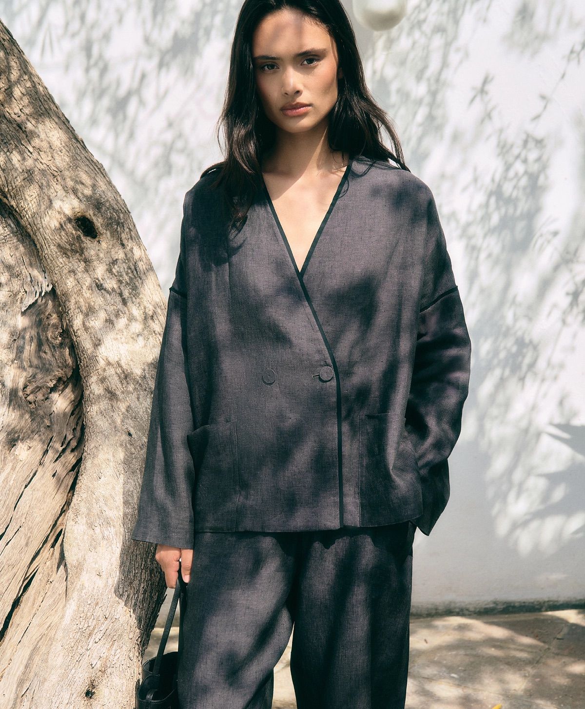 Una donna in tailleur grigio scuro è in piedi accanto a un albero, illuminata da una luce solare filtrata tra gli alberi.