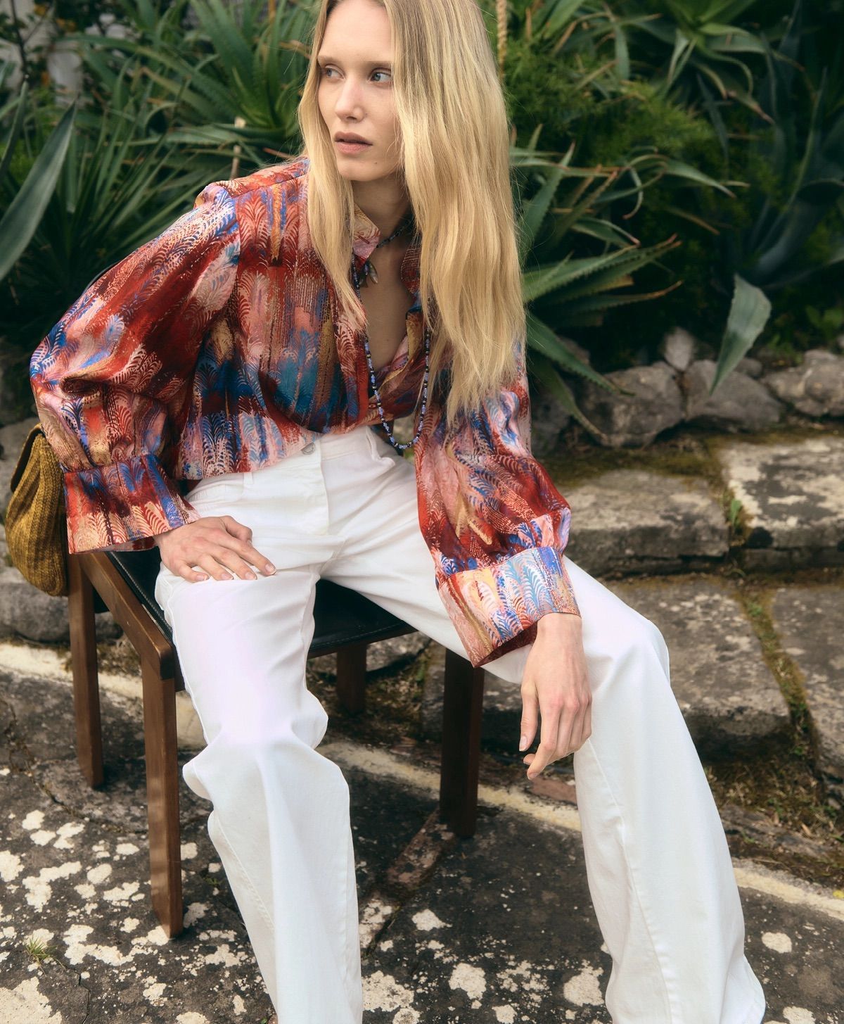 Donna seduta all'aperto con una blusa colorata e pantaloni bianchi, con vegetazione e pietre alle sue spalle.