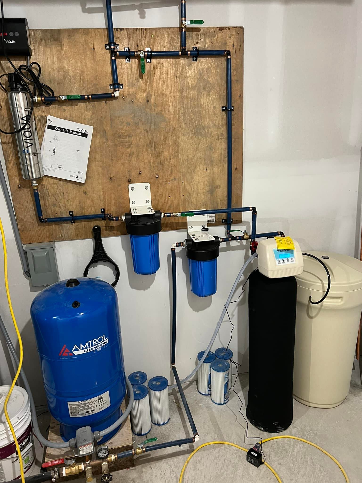 Water filtration system with blue, black, and white components mounted on a wood board in a utility room. Includes filters, a pressure tank, and a water softener.