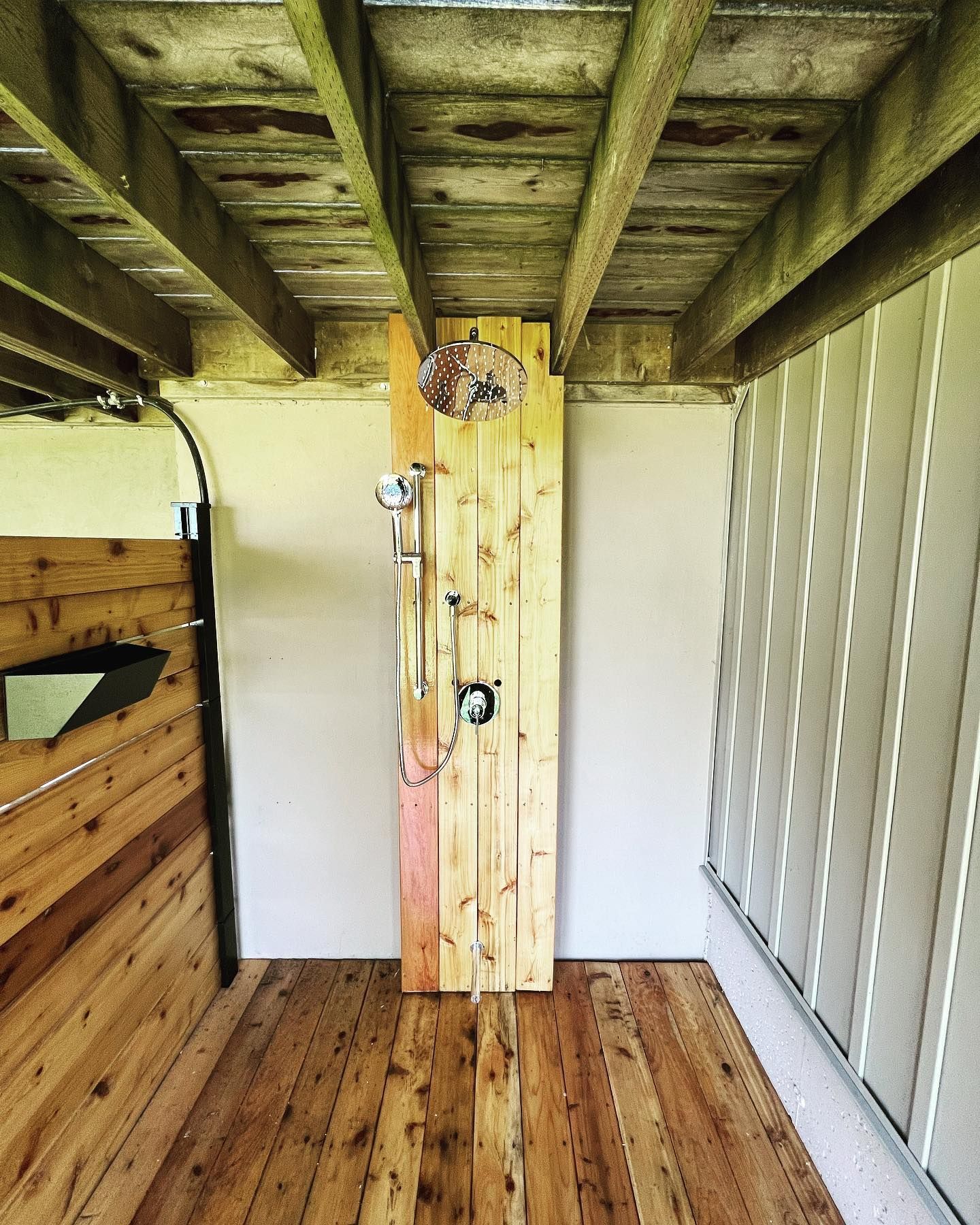 Outdoor shower with wooden plank walls and flooring, under a wooden ceiling. Silver shower fixtures are mounted on a vertical wooden plank.