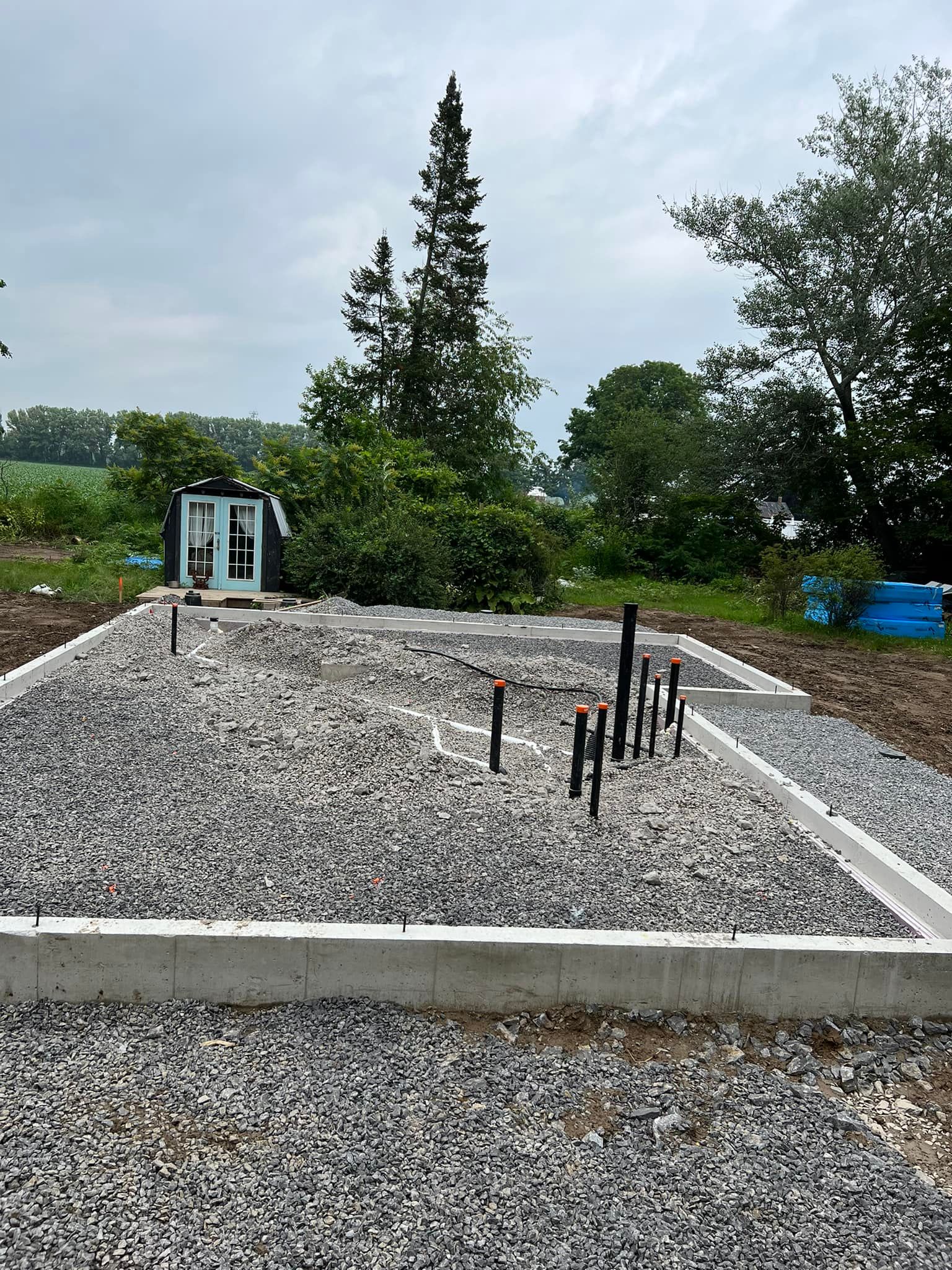 Construction site with concrete foundation, gravel fill, and plumbing pipes. A small structure and trees are in the background under a cloudy sky.