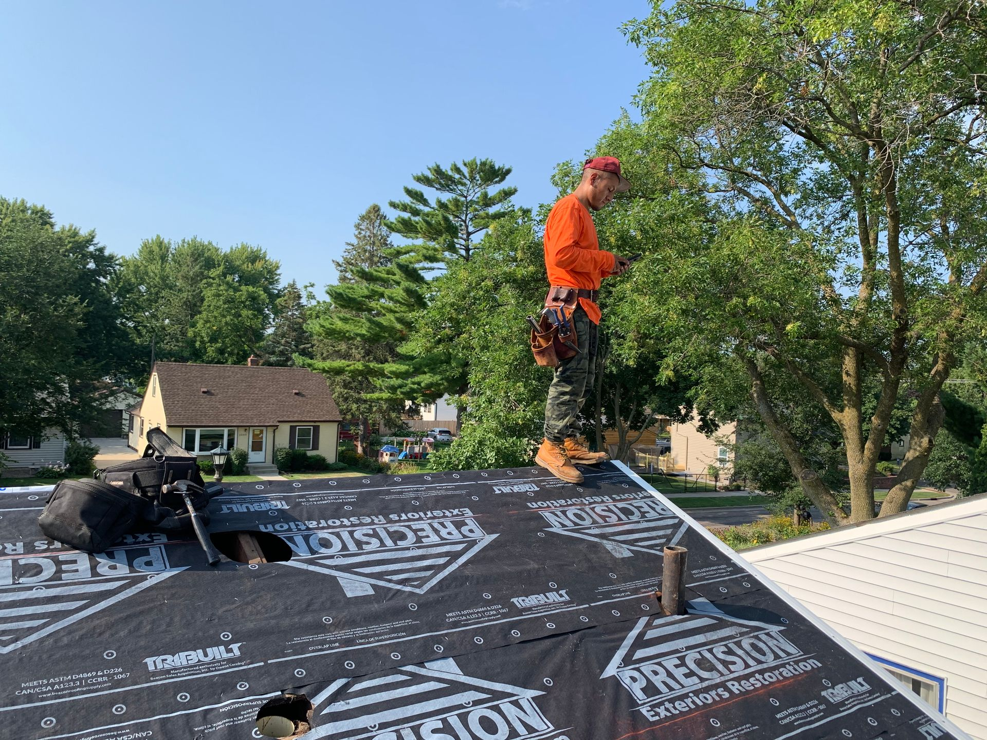 Roofing contractor from Precision Exteriors Restoration standing on a roof underlay, using a smartphone, surrounded by trees and a residential neighborhood in Denver.