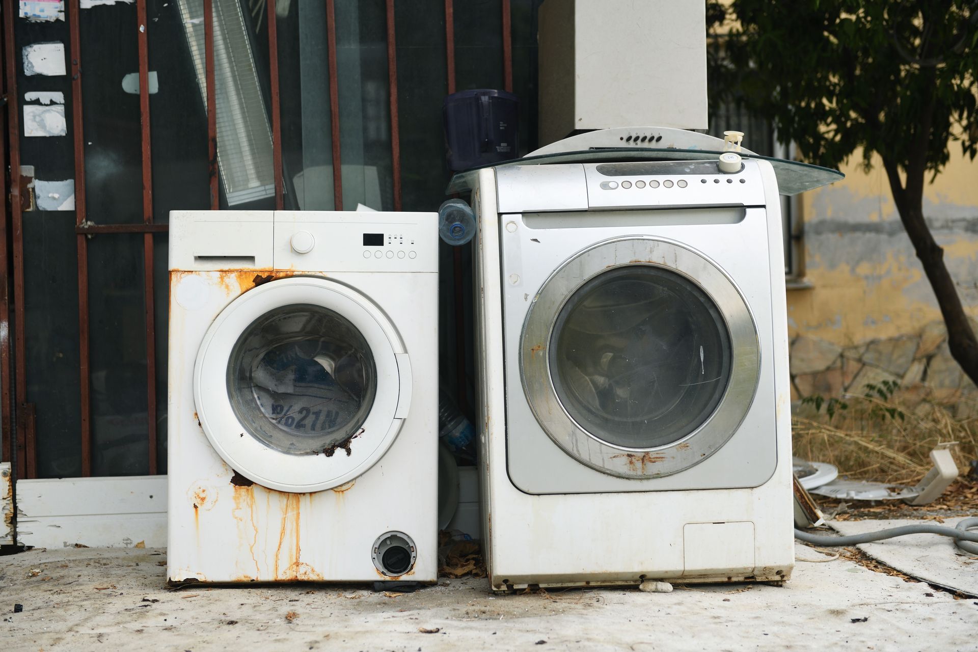 Two old white front-loading washers, one rust-stained, sit outside a building.