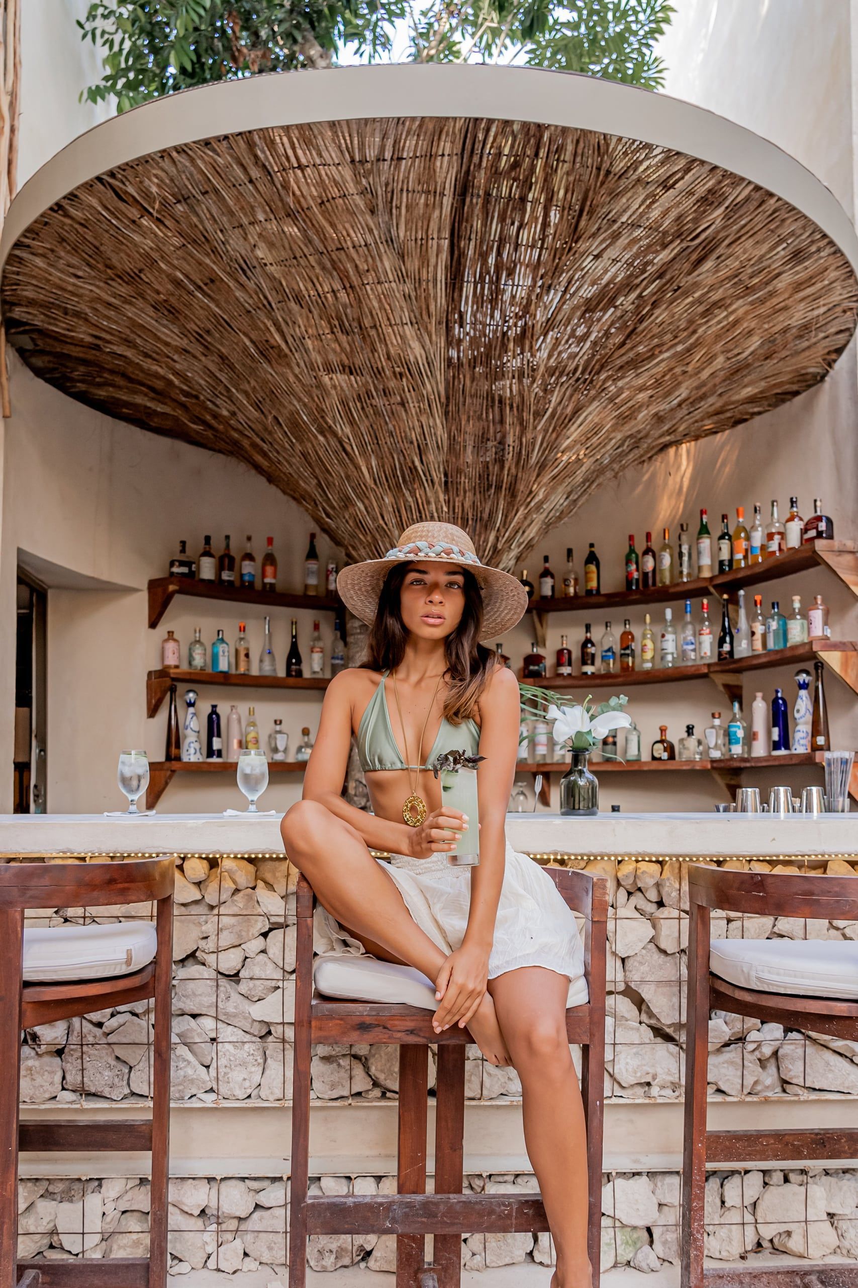 Woman seated at a bar under a large woven canopy, with shelves of bottles behind her