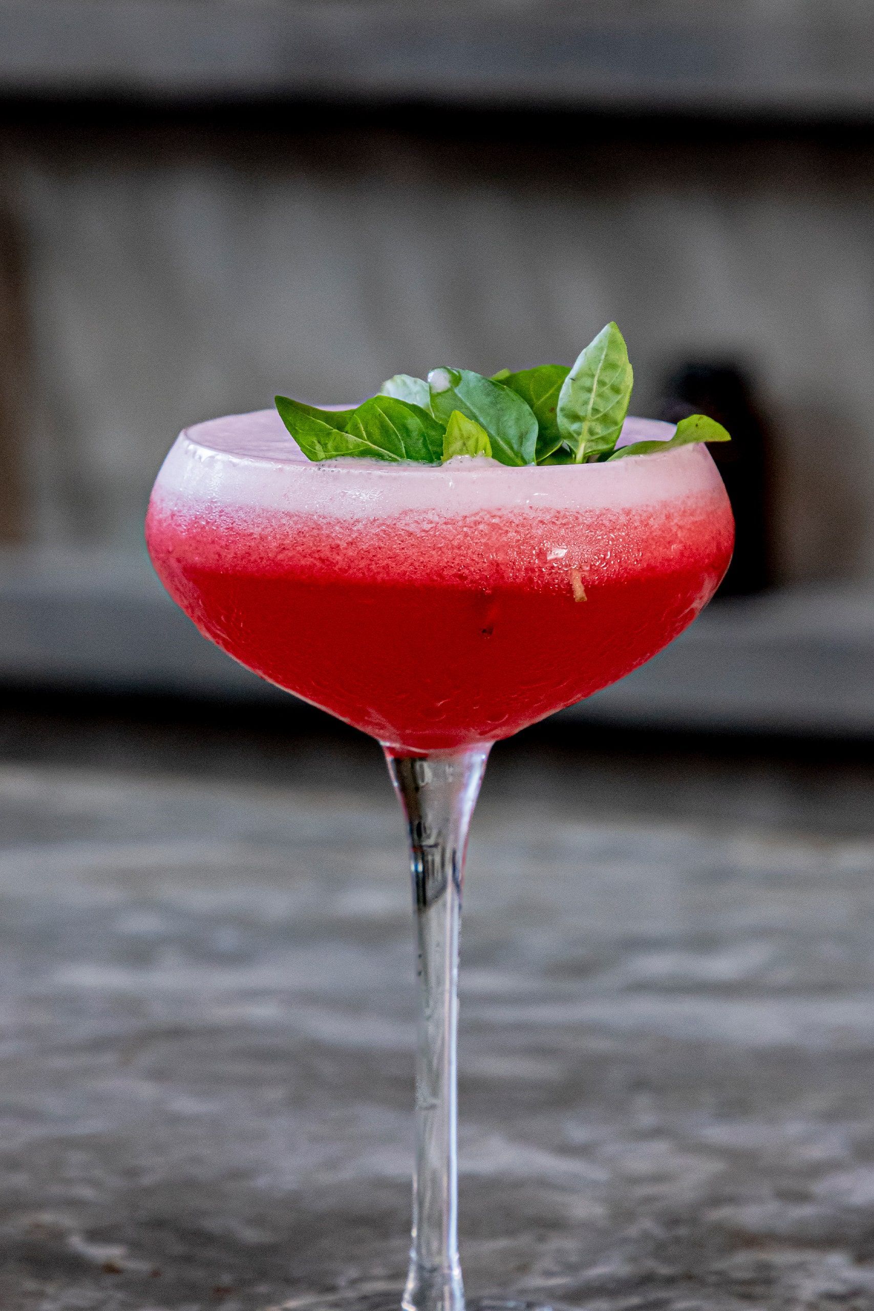 A bright red cocktail in a stemmed coupe glass, topped with fresh green basil leaves, set on a dark textured surface.