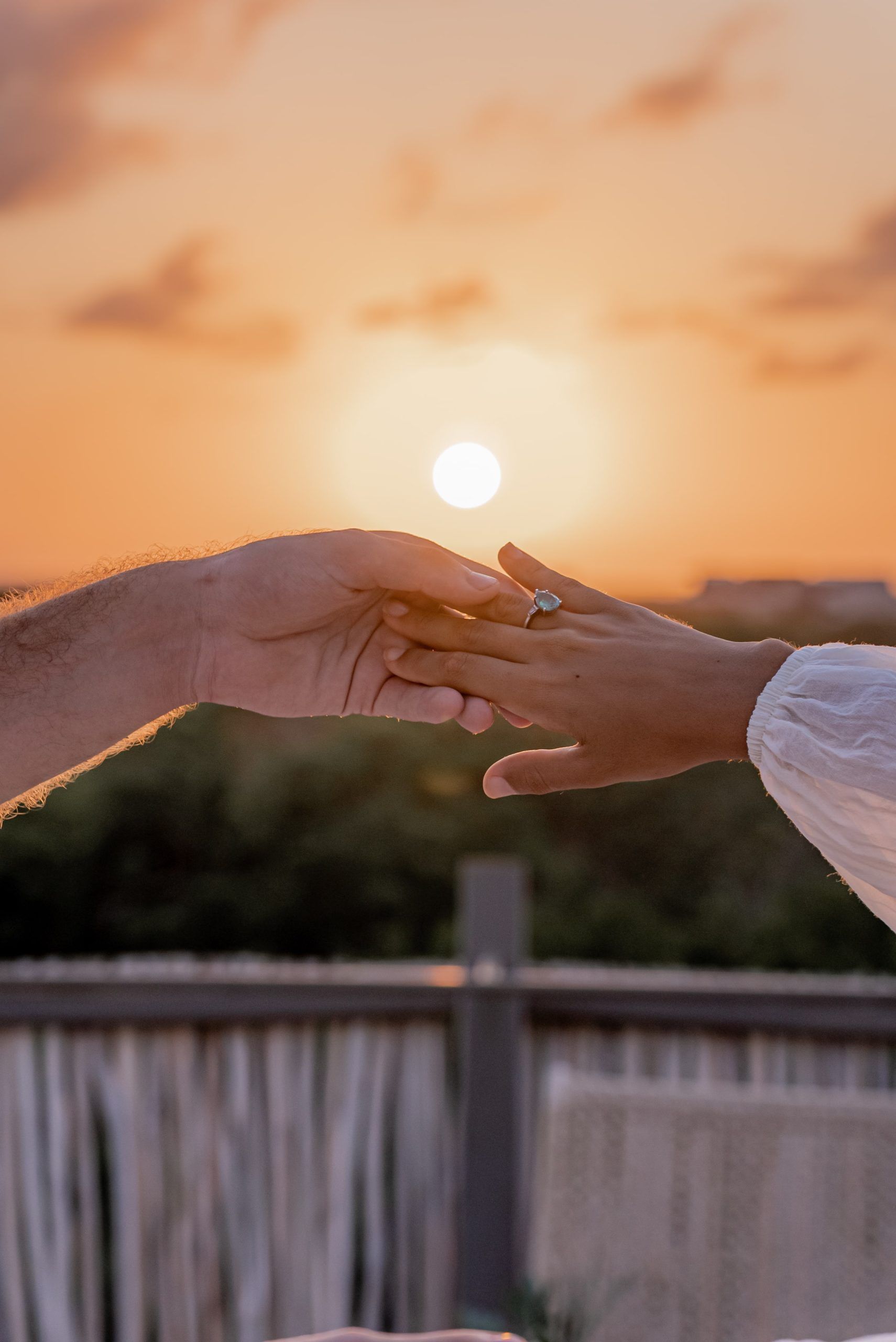 Two hands reach toward each other against a bright, golden sunset over a wooden railing and lush greenery.