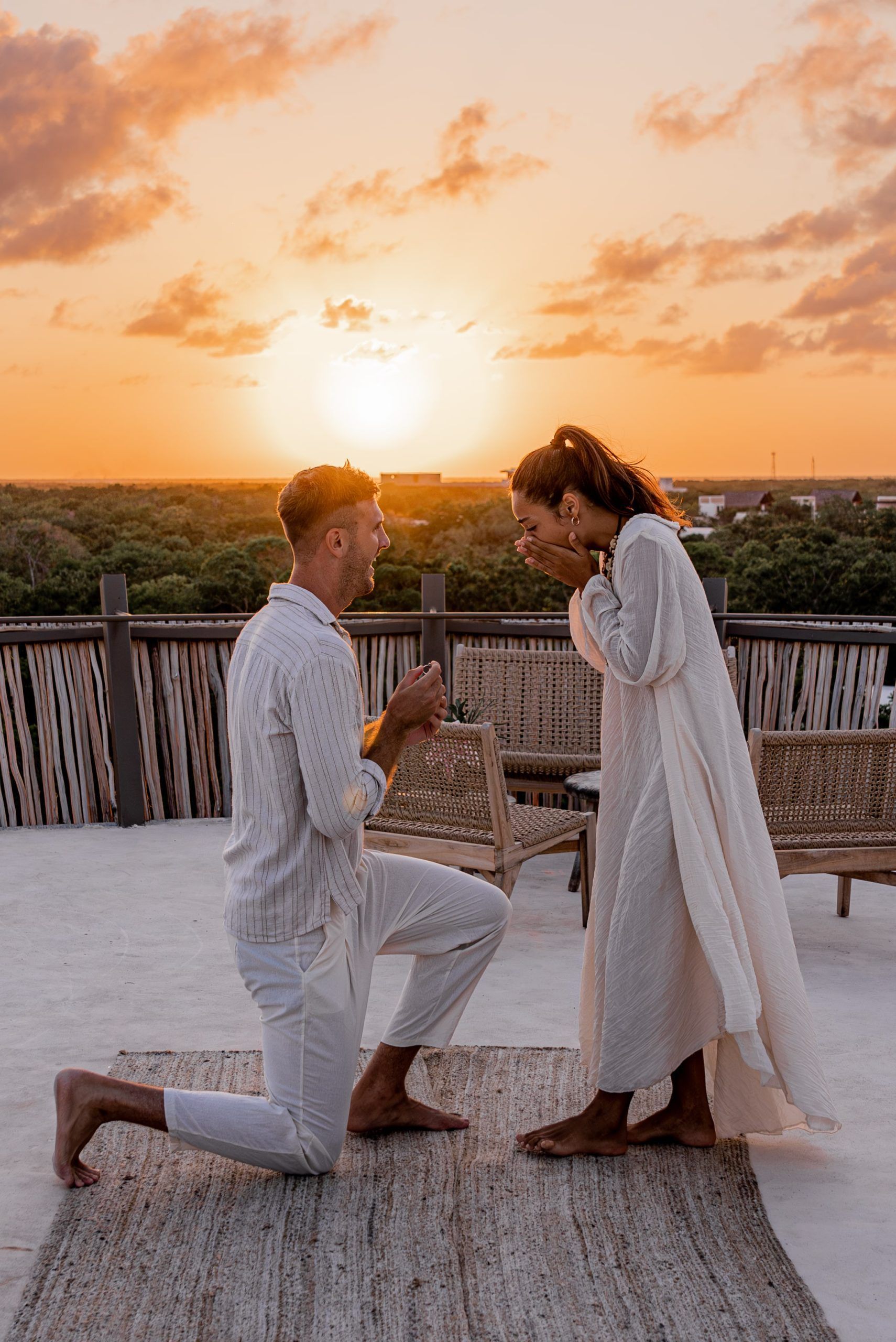 A person kneels to propose to another on a rooftop at sunset, both dressed in white, with a scenic forest backdrop.