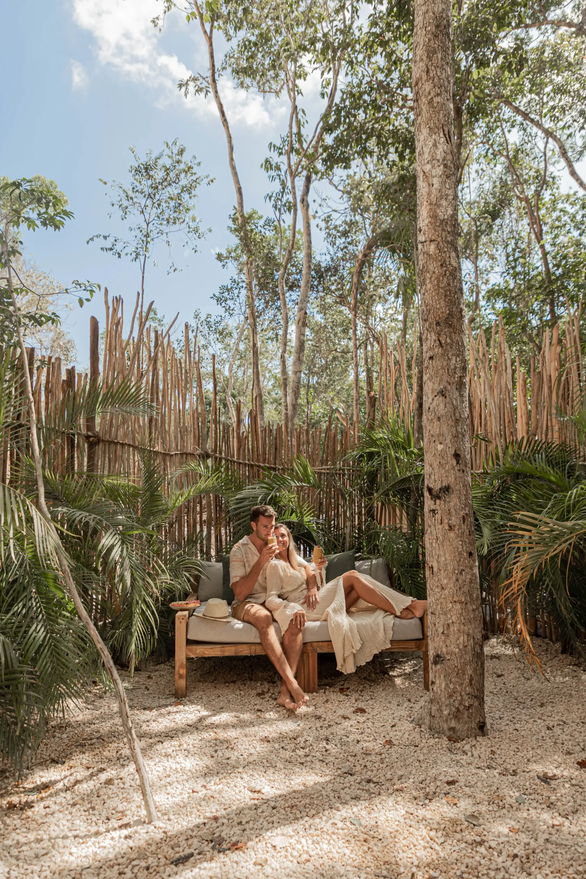 A couple in white robes sits on an outdoor wooden bench surrounded by lush greenery and a tall bamboo fence.