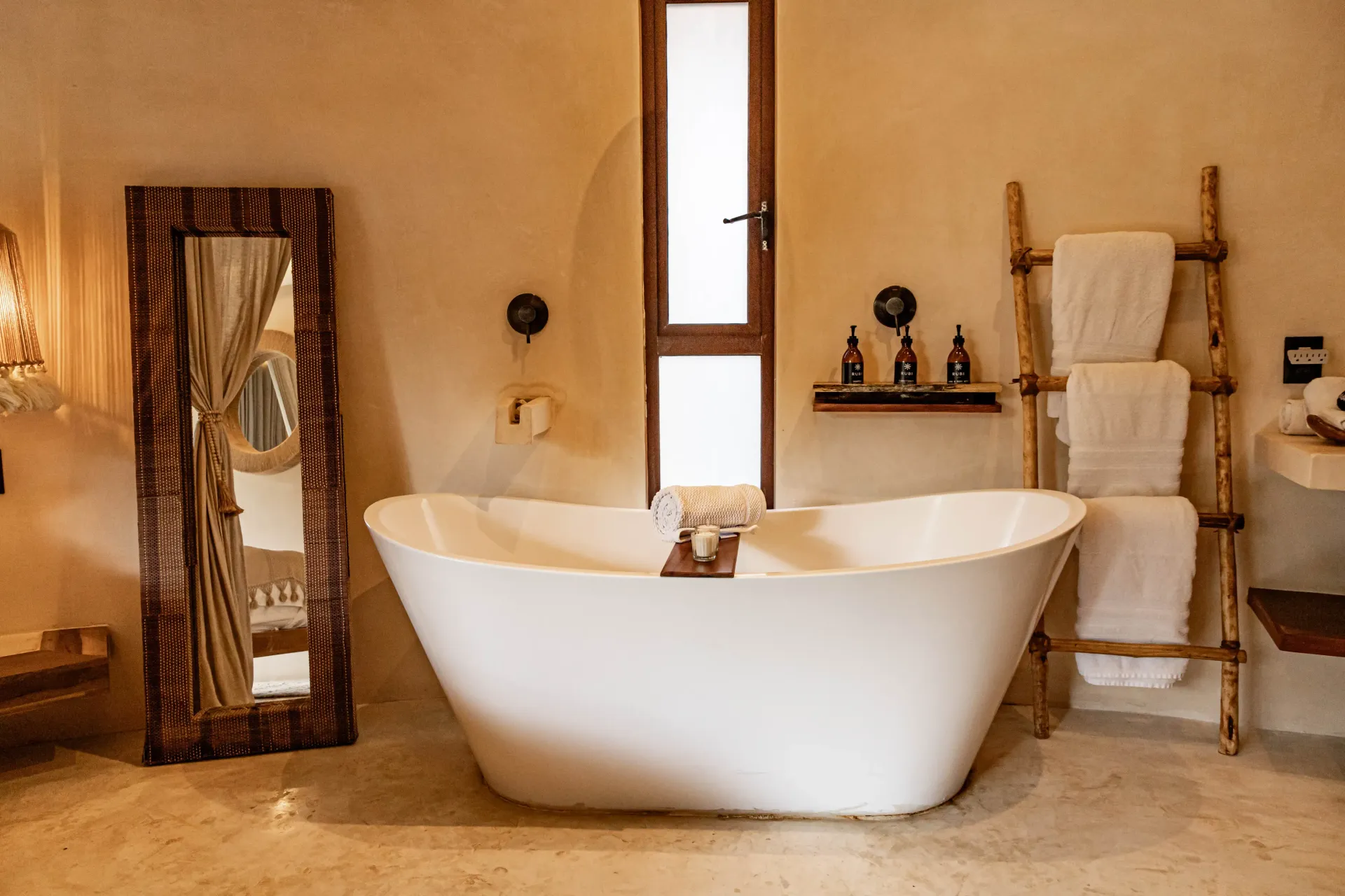 A white standalone bathtub sits in a rustic bathroom with earthy walls, a wooden ladder towel rack, and a floor mirror.