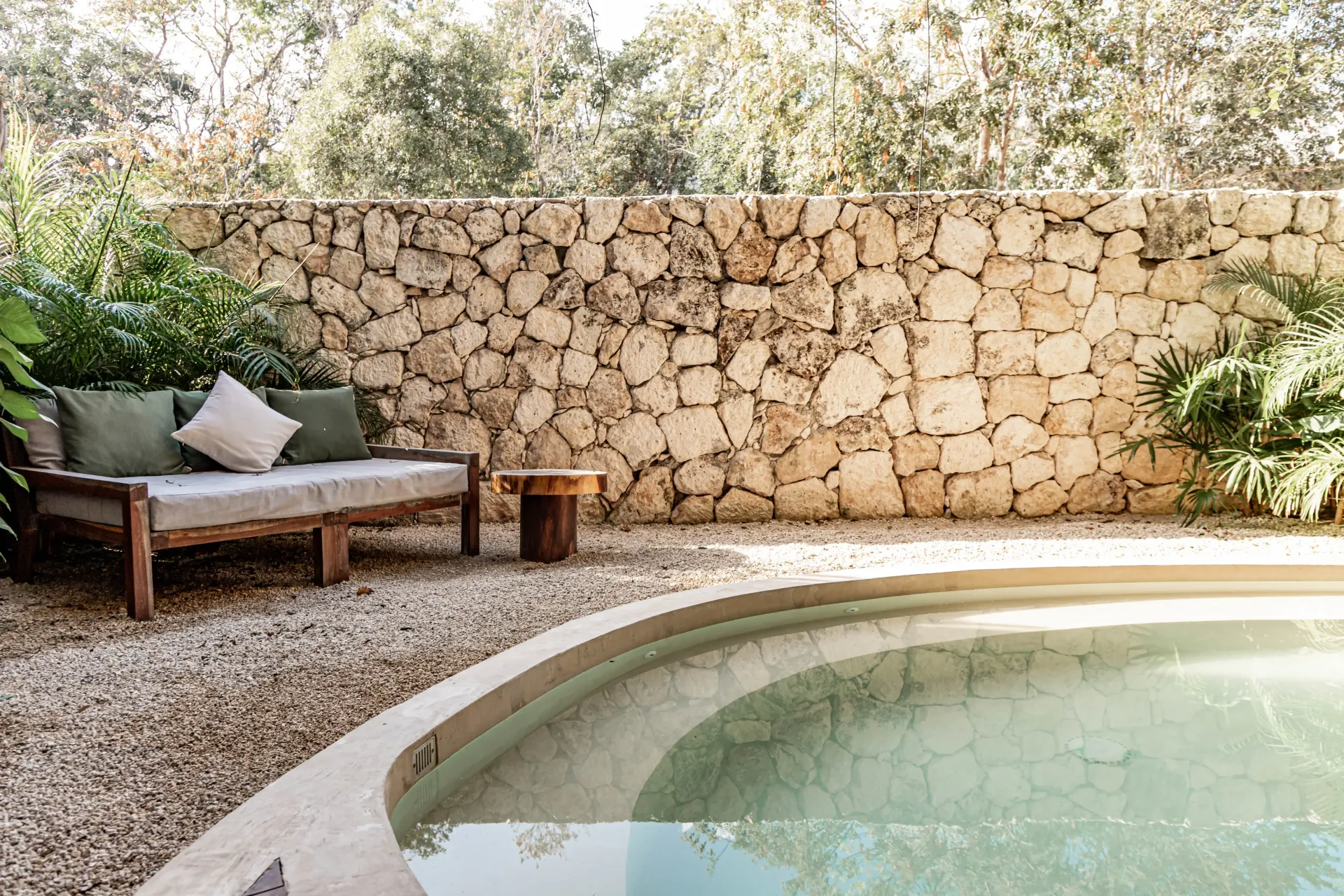 A round pool sits beside a wooden bench with green cushions and a small side table in front of a rustic stone wall.