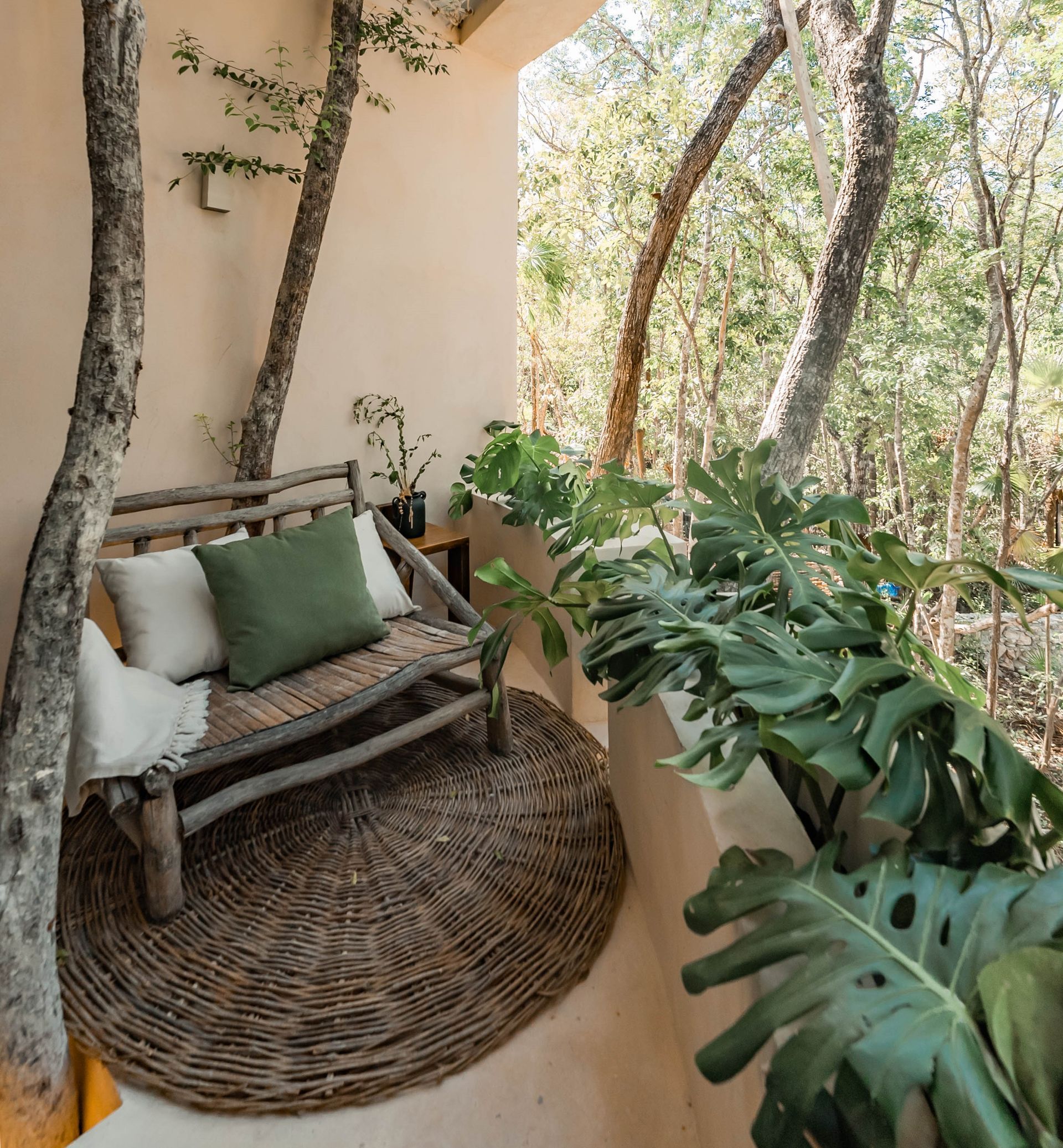 Rustic porch with a woven bench, green cushions, and a round rug beside lush trees and plants.