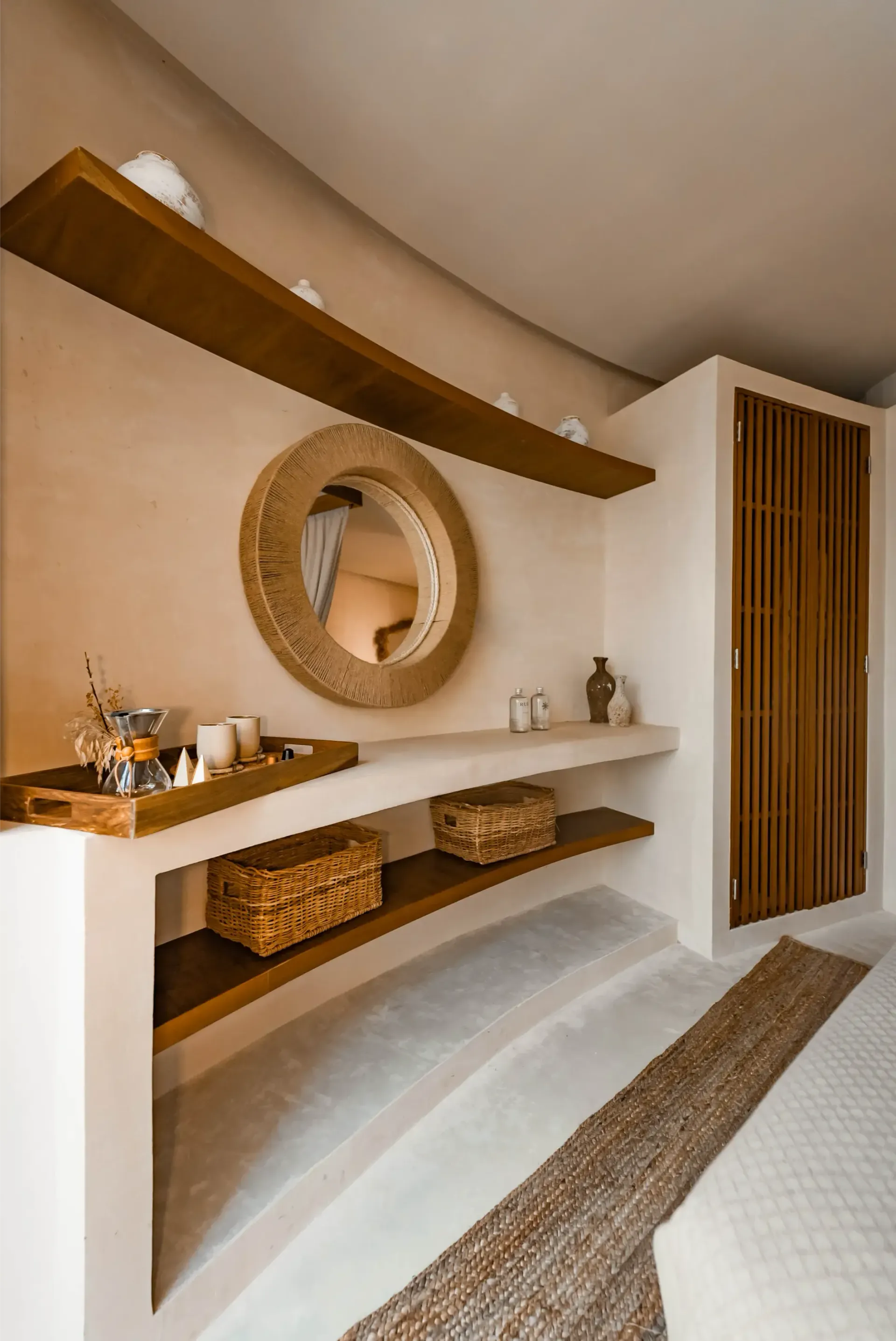 A minimalist bathroom vanity area with a round woven mirror, wooden shelving, and two baskets on a beige plaster wall.